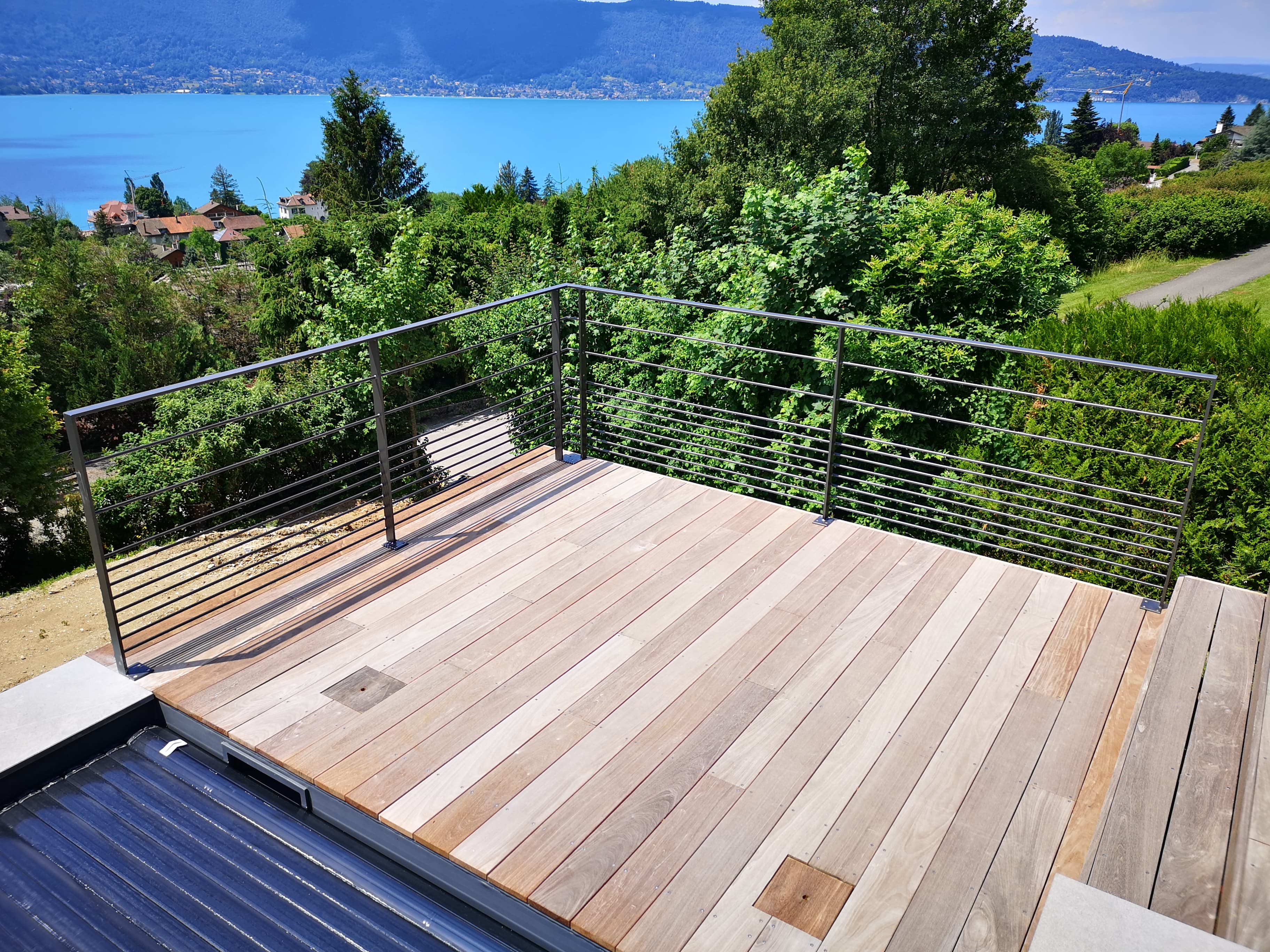 Terrasse en bois avec garde-corps métallique surplombant un lac bleu et des montagnes verdoyantes.