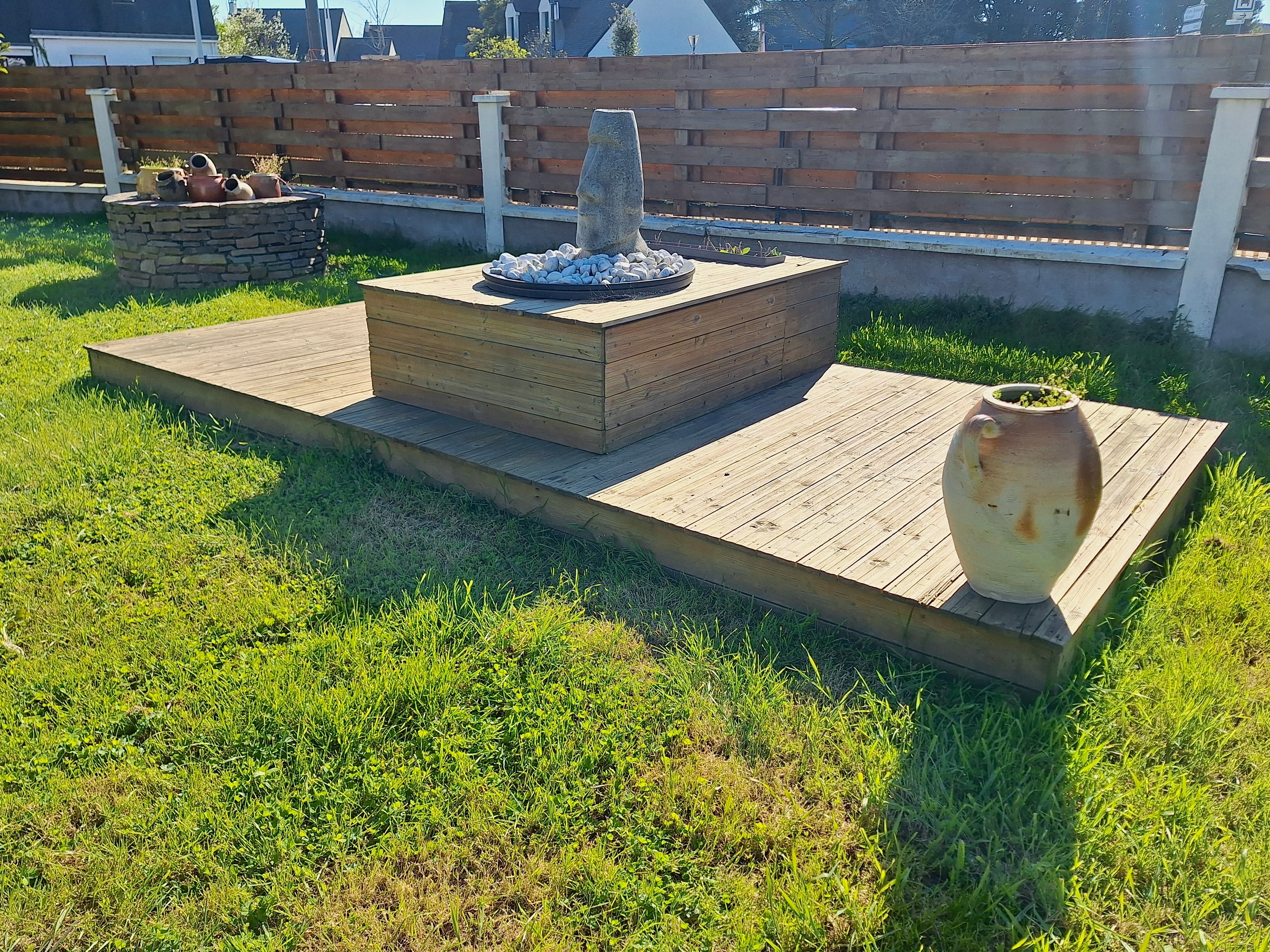 Statue Moai sur terrasse en bois avec jarre en terre cuite dans un jardin ensoleillé.
