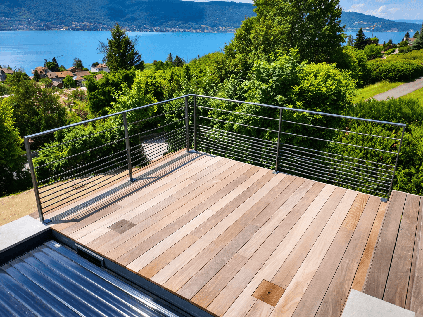 Terrasse en bois avec garde-corps en métal surplombant un lac bleu et des montagnes boisées.